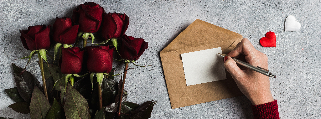 On the right, a person's hand holds a pen to paper atop an envelope, writing a love letter. A bouquet of flowers lays on the left.